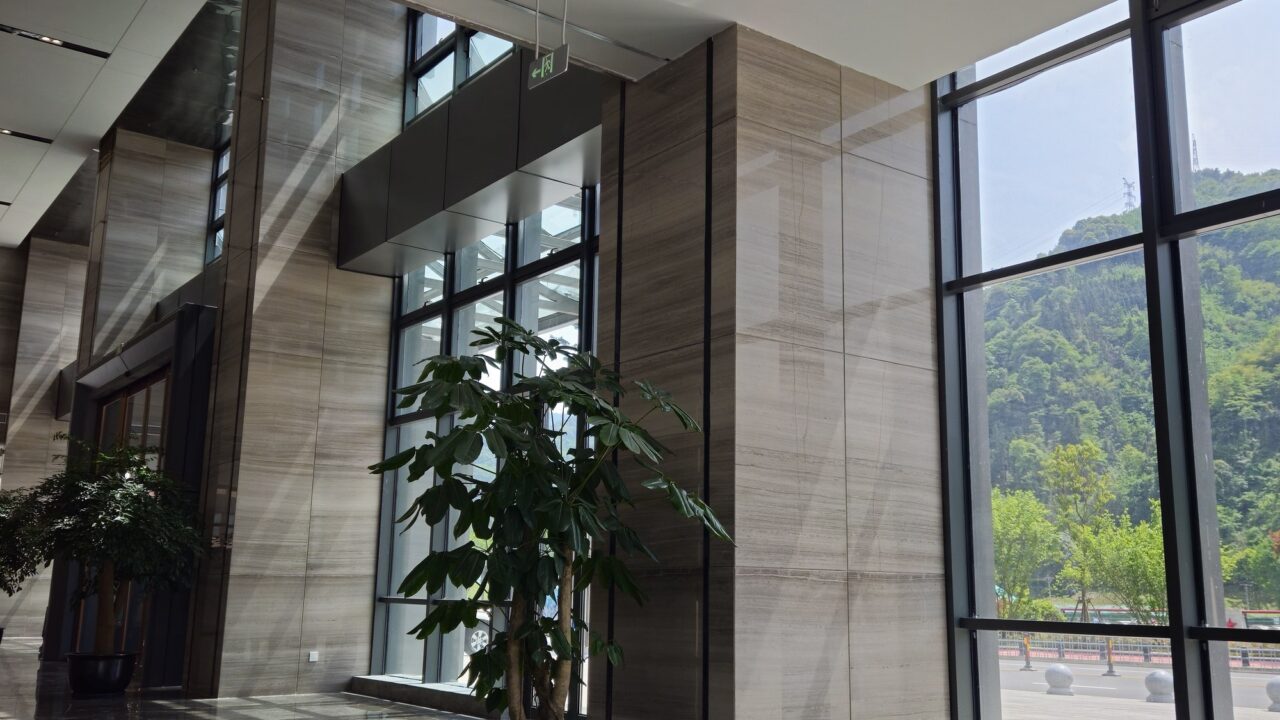 A hotel lobby with floor-to-ceiling windows looking out on a green hillside. Freestanding stone columns and wall panels in a pale grey-brown with straight horizontal bands run the full height of the room, their joints stacked vertically. A potted plant stands in front of them and the floor is a dark grey marble with a white web of veins, with the window frames casting shadows across it.