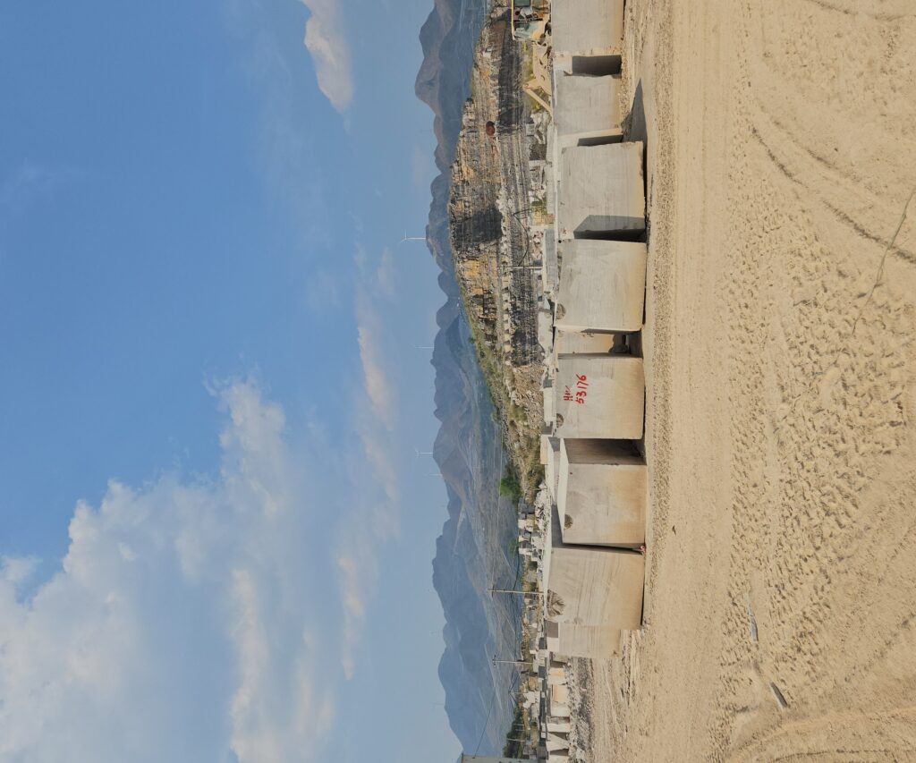 A row of roughly six squared stone blocks standing in the sand at the foot of a quarry, each one cut flat and marked with a red number on its face. Behind them the quarry is cut into benches up the hillside, and wind turbines stand on the ridge above under a blue sky.