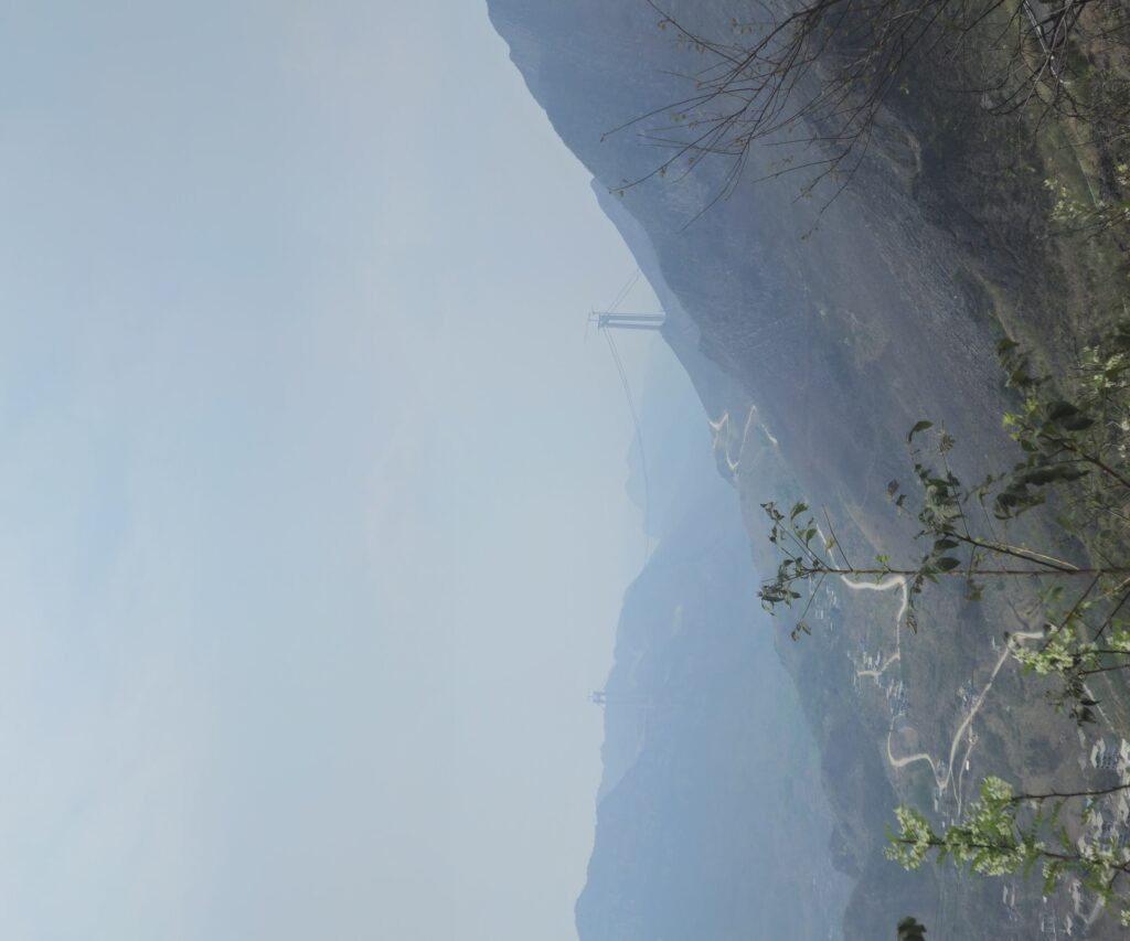 A wide mountain valley seen from high on the slope. Two cableway towers carry a cable across the ridge, a narrow road zig-zags down the mountainside in long bends, and a village lies spread along the valley floor. Bare branches cross the foreground.