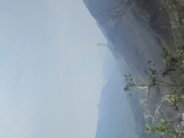 A wide mountain valley seen from high on the slope. Two cableway towers carry a cable across the ridge, a narrow road zig-zags down the mountainside in long bends, and a village lies spread along the valley floor. Bare branches cross the foreground.