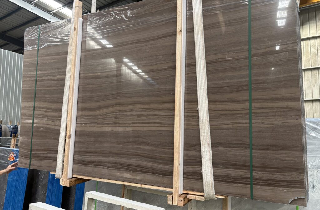 Three dark stone slabs standing upright on timber frames, banded with green straps and wrapped in clear plastic. The faces are a deep warm brown with long parallel waves of figure running across them, darker and more contrasted than the pale boards.