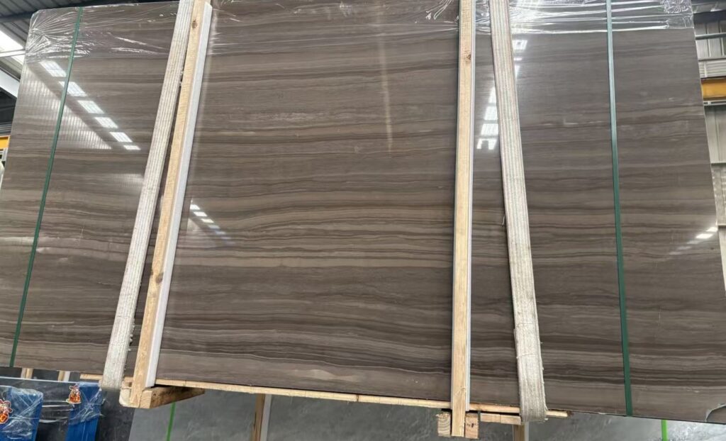 Three stone slabs standing on timber frames, banded with green straps and wrapped in clear film. The faces are a mid warm brown with close parallel lines of figure running across them and a few lighter cloudy patches where the veins spread.