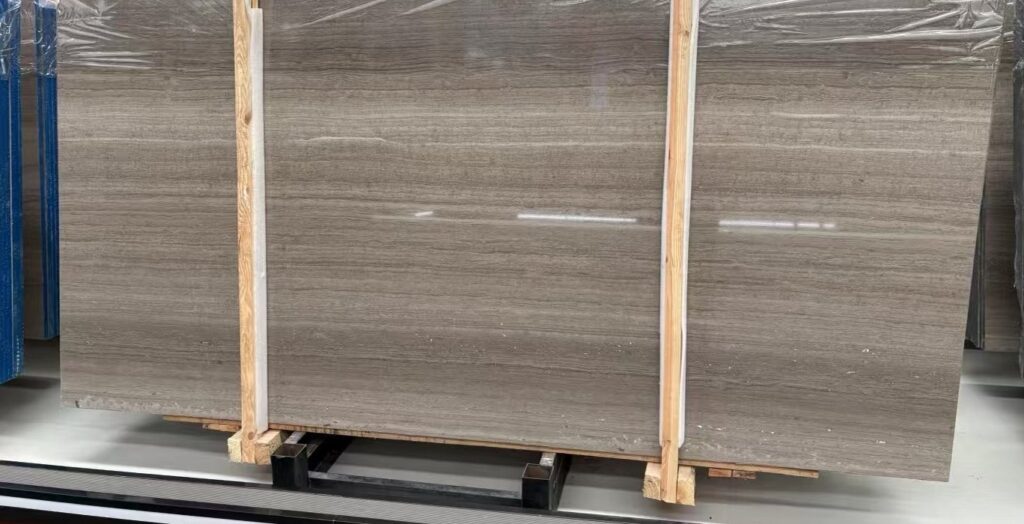 Three pale polished stone slabs standing on timber frames in a workshop. The figure is in fine, closely spaced straight lines, and the polished surface carries a soft reflection of the roof lights along its top edge.