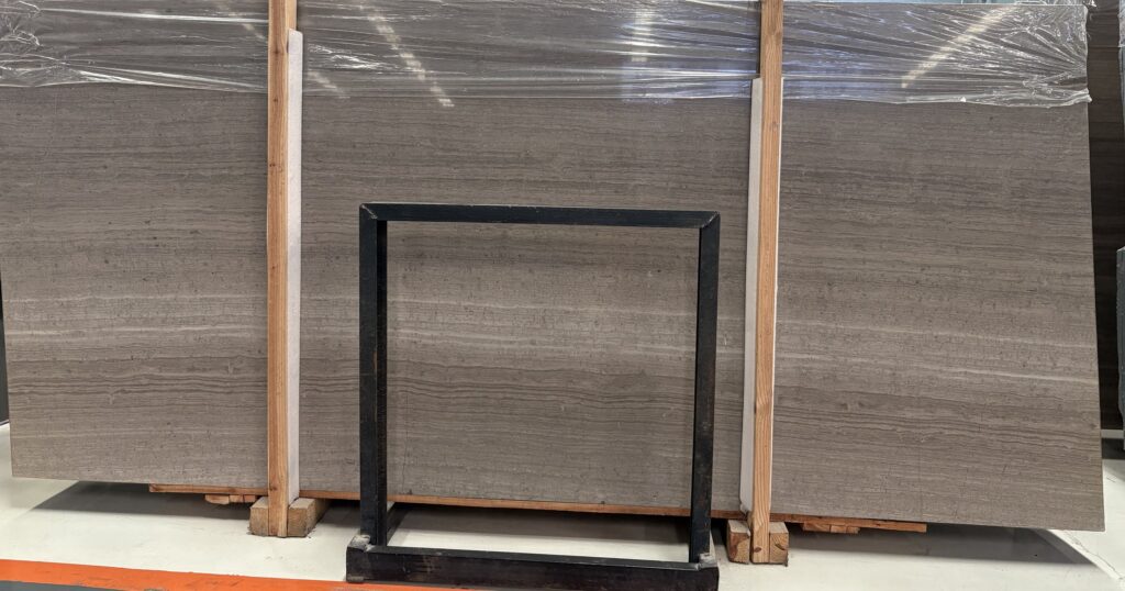 Three polished stone slabs standing in timber frames with a black steel square frame in front of the centre board for scale. The stone is a warm grey-brown with even parallel lines running the width of the board, broken by a few darker knotty patches.