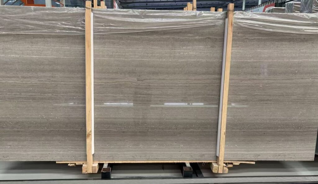 Three polished stone slabs standing upright in timber frames, wrapped in clear plastic, side by side in a workshop. The faces are a flat grey-brown with fine straight parallel lines running across them, and the overhead lights reflect in long streaks along the polish.