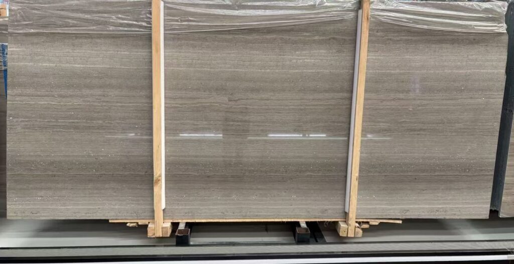 Three polished stone slabs standing side by side in a workshop, wrapped in clear film. These are the flattest and palest faces of the set, with the figure in fine straight lines that barely move across the board.