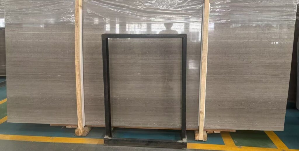 Three polished stone slabs standing upright in timber frames with a black steel square frame in front of the middle one, used to measure the board in the photograph. The faces are a warm grey-brown with evenly spaced straight lines, polished to a mirror finish.
