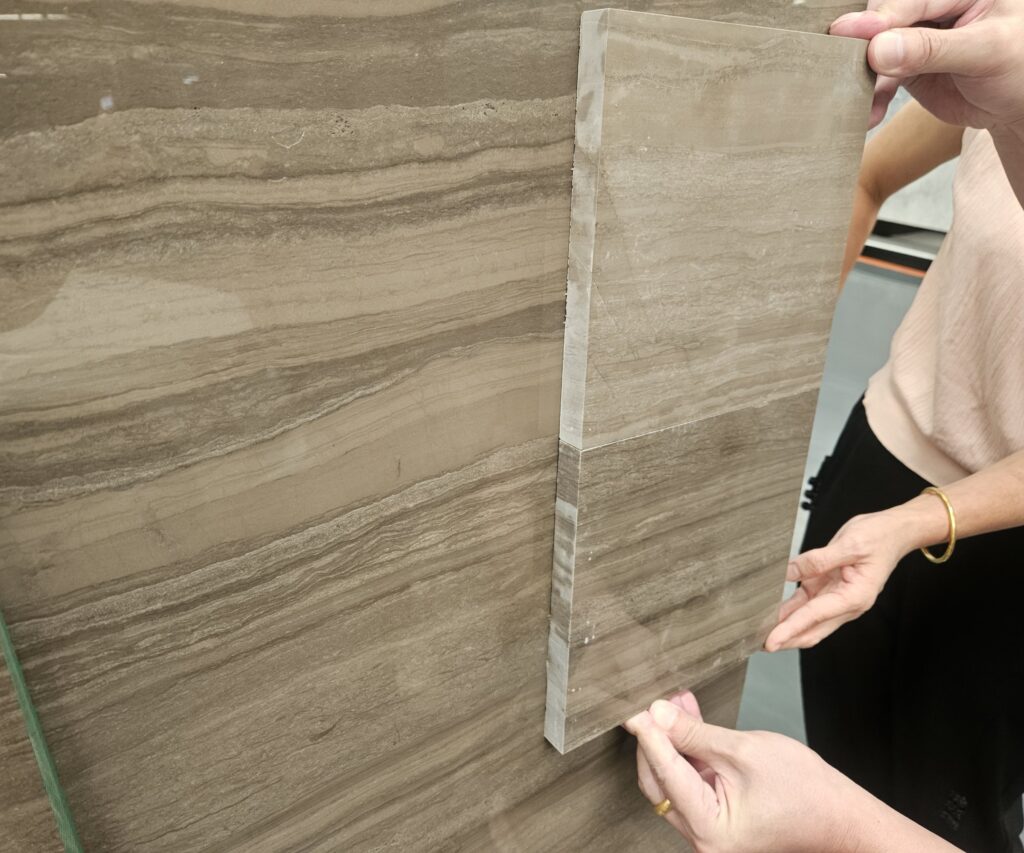 Hands holding a rectangular cut stone sample with a finished edge against the face of a large polished marble slab