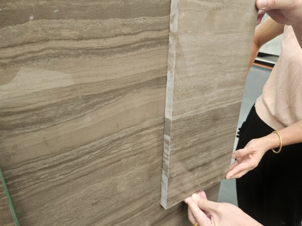 Hands holding a rectangular cut stone sample with a finished edge against the face of a large polished marble slab