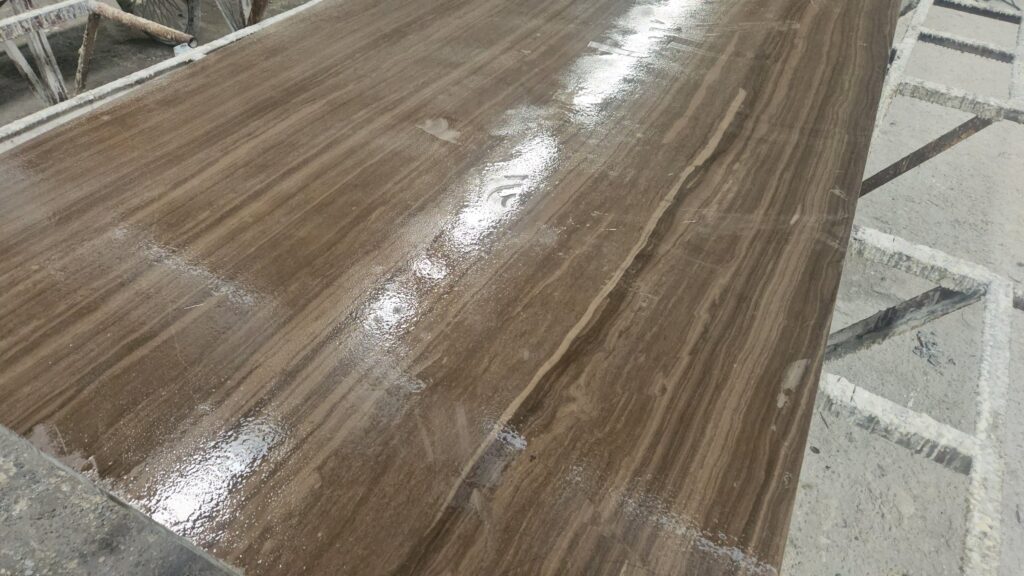 A close view of a wet polished marble slab showing straight dark brown veins running across a warm grey-brown base in even parallel lines