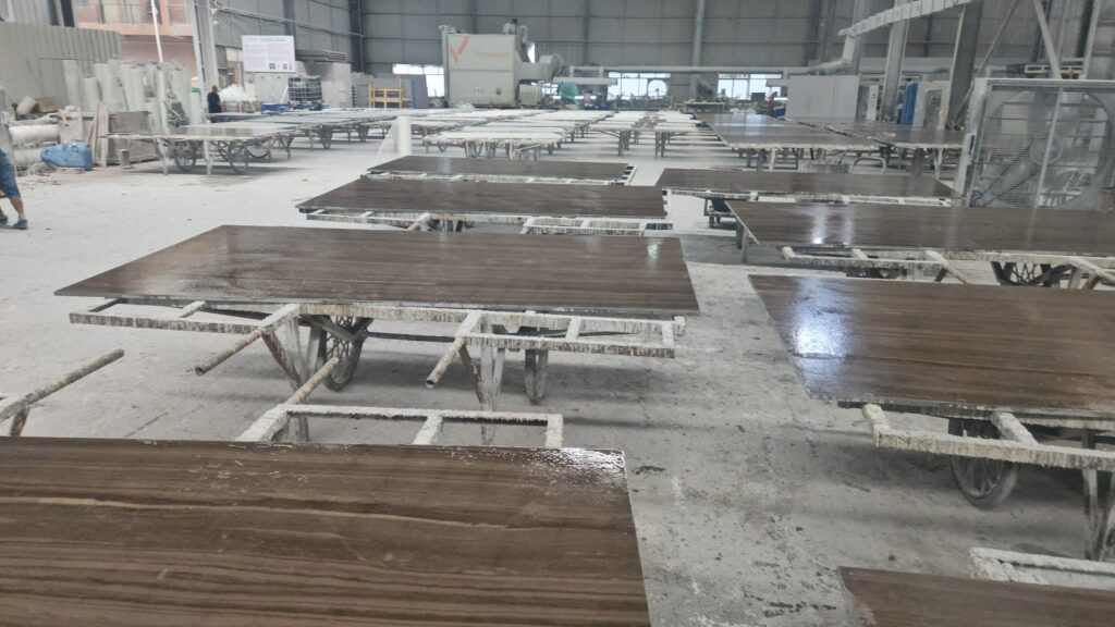 Rows of polished grey-brown marble slabs with straight wood-like grain laid flat on trestles across a large workshop floor, the surfaces wet and reflecting overhead lights